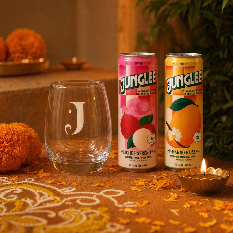 The engraved Junglee J glass beside Lychee Serenity and Mango Bliss cans with a diya and marigold petals